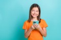 Photo of cheerful small girl wear stylish orange t-shirt hold smartphone look at sale empty space isolated on teal color background.