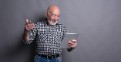 Smiling senior man reading news on digital tablet. Cheerful excited mature male using portable computer, gray studio background